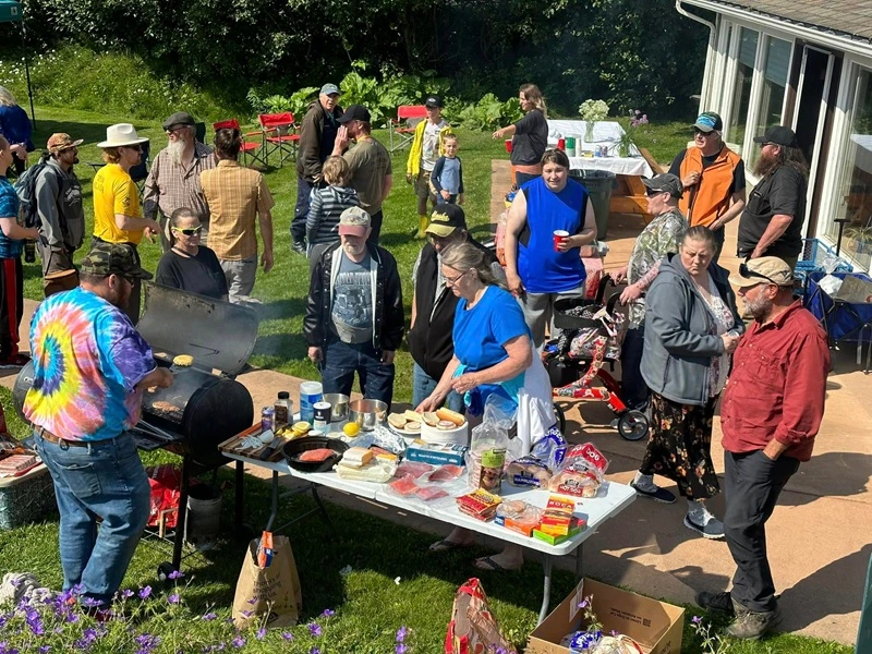 Large group of people gathered outdoors, enjoying a barbecue with food on tables and a grill in the foreground.