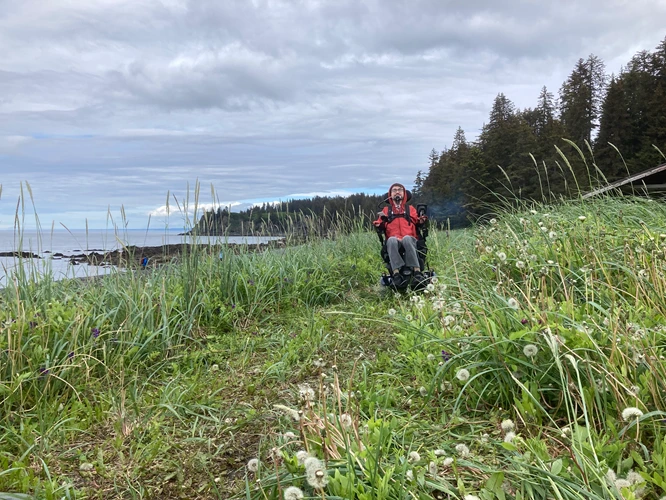 A man in a wheelchair rolling down an overgrown grassy path overlooking the ocean.