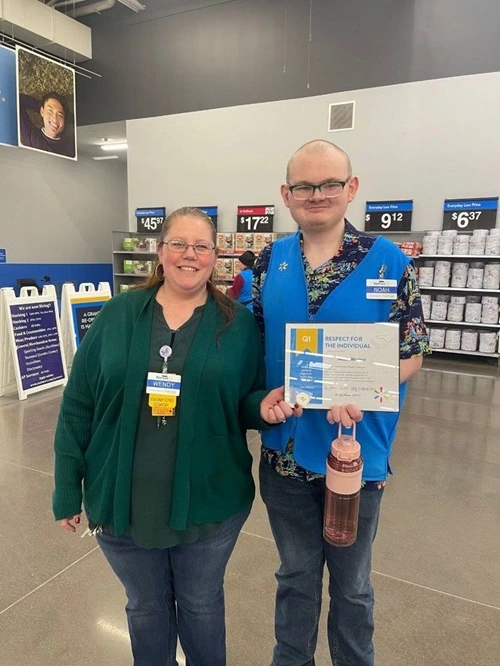 Two employees in a Wal-Mart, wearing nametags reading 'Wendy' and 'Noah'. Noah is holding a certificate that reads 'Respect for the Individual'.