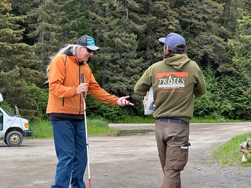 Two people outdoors, passing each other on a dirt road. One of them is holding a white cane, and the other is wearing a sweatshirt that reads 'TRAILS' on the back.